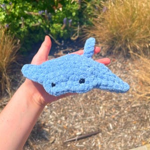 May include: A light blue crocheted manta ray plush toy with black eyes. The handmade sea creature has a textured surface and is held in a person's hand. The background features green foliage and dry grass.