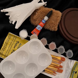 May include: A collection of art supplies including a white bat-shaped sculpture, a brown fuzzy pad, a tube of super glue, a white paint palette, small paint pots, and a set of artist brushes. The items are arranged on a black surface.