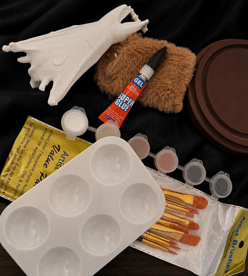 May include: A collection of art supplies including a white bat-shaped sculpture, a brown fuzzy pad, a tube of super glue, a white paint palette, small paint pots, and a set of artist brushes. The items are arranged on a black surface.