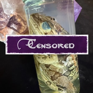 Może przedstawiać: Szklany słoik zawierający zakonserwowaną żabę, ze słowem "Censored" na białym tle na fioletowym banerze. Żaba jest brązowo-zielona, z niebieskim okiem. Słoik jest wypełniony przezroczystym płynem.