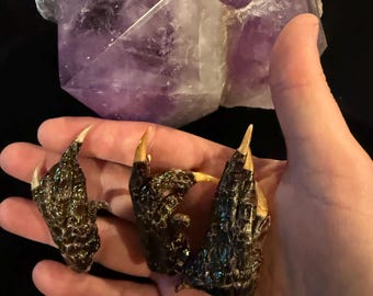 Puede incluir: Tres objetos oscuros en forma de garra, con puntas doradas, se sostienen en una mano. Las garras tienen una apariencia texturizada y escamosa. Una gran formación de cristal púrpura translúcido está en el fondo.