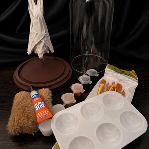 May include: A craft kit with a white sculpture, a glass dome, paint pots, paint brushes, a paint palette, and a tube of Super Glue. The kit includes a brown base and a small, furry item.