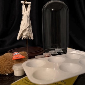 May include: A white bat sculpture hangs from a gold stand, next to a glass cloche. A paint palette, paint tubes, and small jars are also visible. The items are arranged on a wooden base, suggesting a craft or art project.