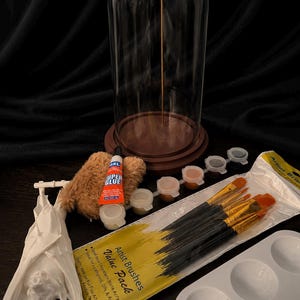 May include: A clear glass cloche with a wooden base and a gold rod inside. The image also shows a white bat figurine, paintbrushes, paint pots, a palette, glue, and a fluffy brown item. The text on the paintbrush package reads "Artist Brushes Value Pack".
