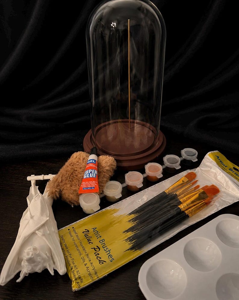 May include: A clear glass cloche with a wooden base and a gold rod inside. The image also shows a white bat figurine, paintbrushes, paint pots, a palette, glue, and a fluffy brown item. The text on the paintbrush package reads "Artist Brushes Value Pack".