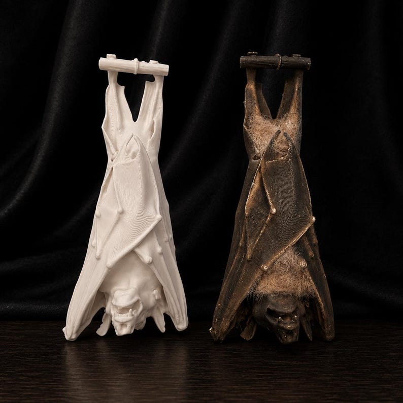 May include: Two bat-shaped sculptures, one white and one brown, hanging upside down. The white bat has a smooth finish, while the brown bat has a textured, aged appearance. Both have detailed facial features and are suspended from a wooden bar.