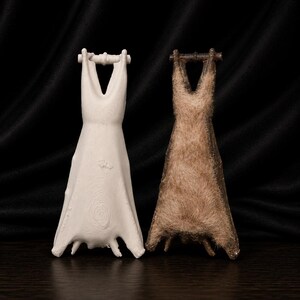 May include: Two decorative sculptures of dresses. One is white and smooth, the other is brown and textured. Both have a V-neck design and are suspended from a horizontal bar. The sculptures are displayed against a dark background.