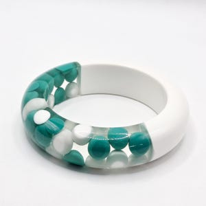 May include: A thick bangle bracelet featuring a clear resin section embedded with numerous small, round, teal and white opaque discs. The remaining portion of the bracelet is constructed from a solid, smooth, opaque white material.