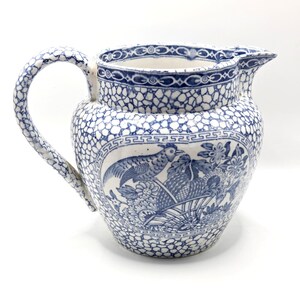 May include: A ceramic pitcher featuring a blue and white transferware pattern. The body displays a central medallion with a bird and fruit basket motif, surrounded by a crackle-like texture and a Greek key border along the rim and handle.