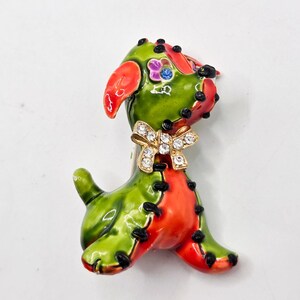 May include: A dog-shaped brooch featuring vibrant green and orange enamel sections separated by black stitch-like detailing. The dog has a small purple flower accent on its head, a blue rhinestone eye, and a gold-tone bow encrusted with clear rhinestones.