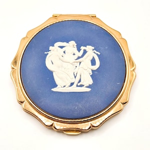 May include: A round gold-tone metal compact mirror featuring a blue circular inlay with a white relief design of three figures dancing. The metal frame has a scalloped edge, and the central blue disc displays a classical scene with draped fabric.