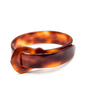 May include: A brown and amber tortoise shell patterned bangle bracelet with a smooth, glossy finish. The bracelet features a buckle-style closure design where one end of the band loops through a fixed, integrated loop on the other side.