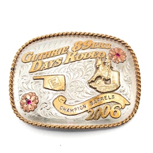 May include: An oval belt buckle with a silver-toned engraved background and gold-toned accents. It features the text GUTHRIE 89ERS DAYS RODEO, a map of Oklahoma, a horse and rider, a banner reading CHAMPION BARRELS, the year 2006, and two floral motifs with red stones.