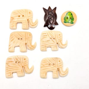 May include: A collection of decorative buttons in various shapes. There are six ivory-colored elephant buttons with etched details, a dark brown bat-shaped button, and a round button with a green frog design.