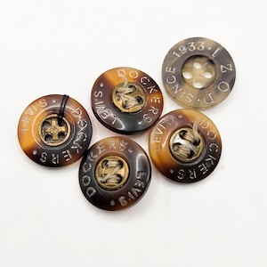May include: A collection of brown and tan Levi's Dockers buttons. The buttons have a circular shape with the words "LEVI'S DOCKERS" and "SINCE 1933-1" etched around the edges. Each button has a metal center.