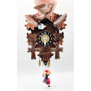 May include: A wooden cuckoo clock with carved leaf and bird details. The clock face is black with white numbers. A small figure swings below the clock. The clock is held by a hand.