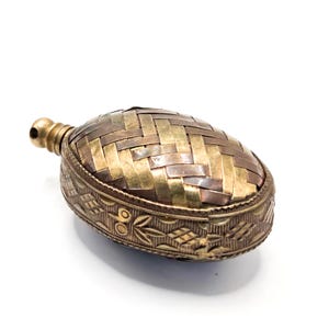 May include: An oval-shaped metal flask featuring a woven pattern of alternating brass and copper-toned strips on the top surface. The sides of the flask are decorated with an engraved floral and geometric motif, and it has a small threaded neck.