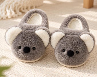 May include: A pair of plush, grey and white koala-themed slippers featuring soft faux fur, rounded ears, black button noses, and small dark eyes. The interior lining is a cream-colored fabric, and the slippers have a rounded, closed-toe design.