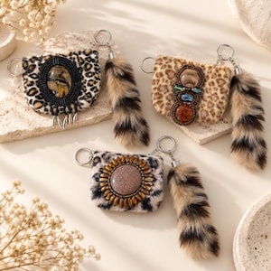 May include: Three small, rectangular, leopard-print faux fur pouches, each featuring a metal keyring and a long, striped faux fur tail charm. Each pouch is decorated with a unique, large, central beaded embellishment in black, brown, or tan tones.