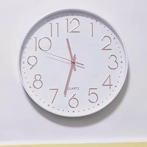 Puede incluir: Un reloj redondo blanco con números romanos negros para las horas y puntos negros para los minutos. Las manecillas del reloj son de oro rosa y la esfera del reloj dice "QUARTZ".