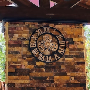 Könnte beinhalten: Eine schwarze und goldene Uhr mit römischen Ziffern und Zahnrädern. Die Uhr ist an einer Steinwand montiert.