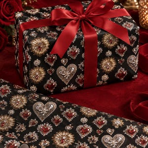 Peut inclure: Un coffret cadeau rectangulaire enveloppé dans du papier noir avec un motif répétitif de soleils dorés et de cœurs ornés de cristaux rouges et transparents, noué avec un large ruban en satin rouge profond formant un nœud sur le dessus.