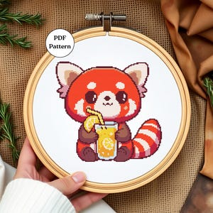 May include: Cross stitch pattern featuring a red panda holding a glass of orange juice with a lemon slice. The pattern is in a wooden embroidery hoop.