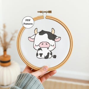 May include: A white and black cow with closed eyes, sitting on a white background. The cow has a pink nose and is surrounded by a wooden embroidery hoop. The text "PDF Pattern" is visible in the top left corner of the image.