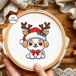 May include: Cross stitch pattern of a cute cartoon dog wearing a Santa hat and reindeer antlers. The dog is wearing a red bow and has a white background.