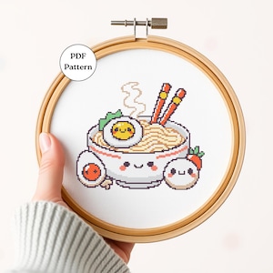 Może przedstawiać: Wzór do haftu krzyżykowego przedstawiający miskę ramen z uśmiechniętą buzią, uśmiechnięte jajko i dwa uśmiechnięte pierożki. Wzór jest wyświetlany w drewnianym kółku do haftu.