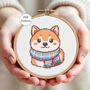 Puede incluir: Un patrón de punto de cruz de un perro Shiba Inu con una bufanda azul, roja y blanca. El perro es naranja y blanco con ojos negros y una nariz negra. El patrón está en un bastidor de bordado de madera. El texto "PDF Pattern" está en un círculo blanco.
