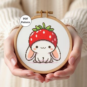 May include: A cross-stitch embroidery hoop showing a cute bunny wearing a strawberry hat. The bunny is white with pink ears and a smiling face. The strawberry hat is red with green leaves. The text "PDF Pattern" is in a white circle.