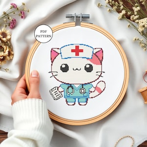 Peut inclure: Modèle de point de croix d'un adorable chat blanc portant un uniforme d'infirmière bleu avec une croix rouge sur son chapeau. Le chat tient un presse-papiers et a un stéthoscope autour du cou. Le motif est affiché sur un cerceau à broder en bois.