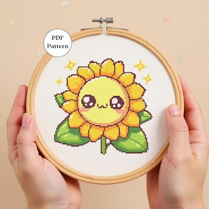 Puede incluir: Patrón de punto de cruz de un girasol sonriente con ojos grandes y pétalos amarillos, hojas verdes y el texto "PDF Pattern" en un círculo blanco. El diseño está en un bastidor de bordado de madera.
