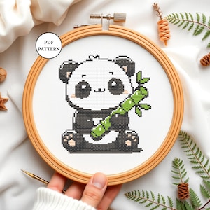 May include: A cross stitch pattern featuring a cute cartoon panda holding a piece of bamboo. The pattern is in a wooden embroidery hoop, with a white background.