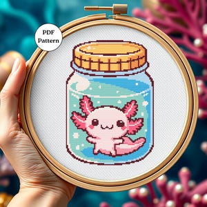 May include: A cross-stitch pattern of a cute axolotl in a glass jar. The axolotl is pink with black eyes and a small smile. The jar is blue with a white lid and has a few bubbles inside. The pattern is in a hoop and is ready to be stitched.