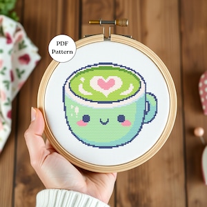 May include: A cross-stitch pattern of a green and blue mug with a smiling face and a heart-shaped design in the green liquid. The pattern is displayed in a wooden embroidery hoop.