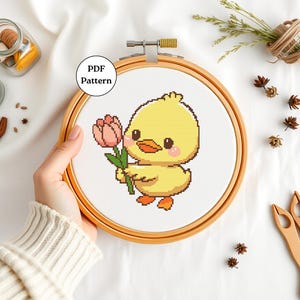Puede incluir: Un pollito amarillo sosteniendo una flor de tulipán rosa en un aro de bordado de madera. El texto "PDF Pattern" es visible en la imagen.