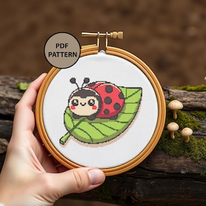 Könnte beinhalten: Ein Kreuzstichmuster mit einer niedlichen Cartoon-Marienkäfer mit schwarzen Punkten auf einem grünen Blatt. Das Muster wird in einem hölzernen Stickrahmen angezeigt. Der Text "PDF PATTERN" ist in der oberen linken Ecke des Bildes sichtbar.