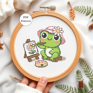 May include: Cross-stitch pattern of a green frog wearing a pink hat with a daisy, painting a flower on a small easel. The design is set in a wooden embroidery hoop. The text "PDF Pattern" is in a white circle.