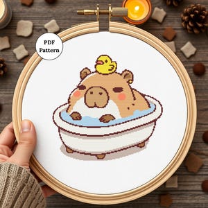 Puede incluir: Patrón de punto de cruz con un capibara marrón con un pato de goma amarillo en la cabeza, relajándose en una bañera blanca llena de agua azul. El patrón se muestra en un aro de bordado de madera.