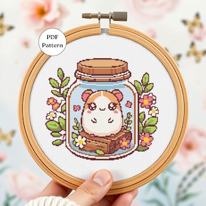 Puede incluir: Un patrón de punto de cruz con un lindo hamster de dibujos animados dentro de un frasco de vidrio con flores y hojas. El frasco es marrón con una tapa blanca. El hámster es marrón y blanco con mejillas rosadas y una nariz rosa. Las flores son rosas y blancas. Las hojas son verdes.