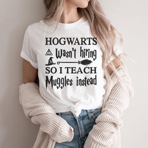 May include: White t-shirt with black text that reads "HOGWARTS Wasn't hiring SO I TEACH Muggles instead." The shirt features a broom, a witch's hat, and a triangle symbol. The shirt is paired with blue jeans and a cream-colored cardigan.