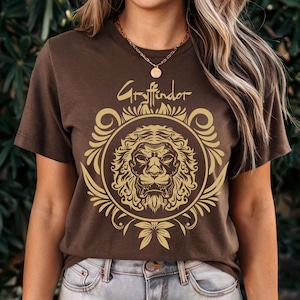 May include: Brown t-shirt featuring a gold lion emblem within a circular frame, with the word "Gryffindor" above. The design includes ornate details and a bow at the bottom. The shirt is a short-sleeved crew neck.