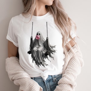 May include: A white short-sleeved t-shirt featuring a graphic print of a ghost sitting on a swing. The ghost is draped in a grey sheet and is blowing a large, round, bright pink bubble with bubblegum. The swing is suspended by black chains.