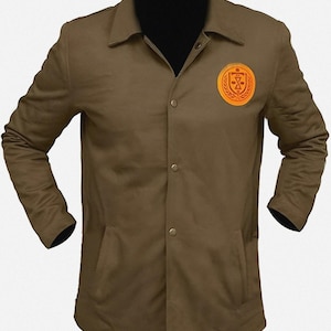 May include: An olive green jacket with a button-up front, collared neckline, and two front pockets. The jacket has a circular emblem on the chest with a gold and orange design. The sleeves have black cuffs.
