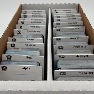 May include: A cardboard storage box filled with stacks of collectible trading cards. Each stack is labeled with the set name, including "Alpha," "Beta," "Unlimited," and various editions of "Magic" cards, such as "Magic 2010" and "Magic 2015."