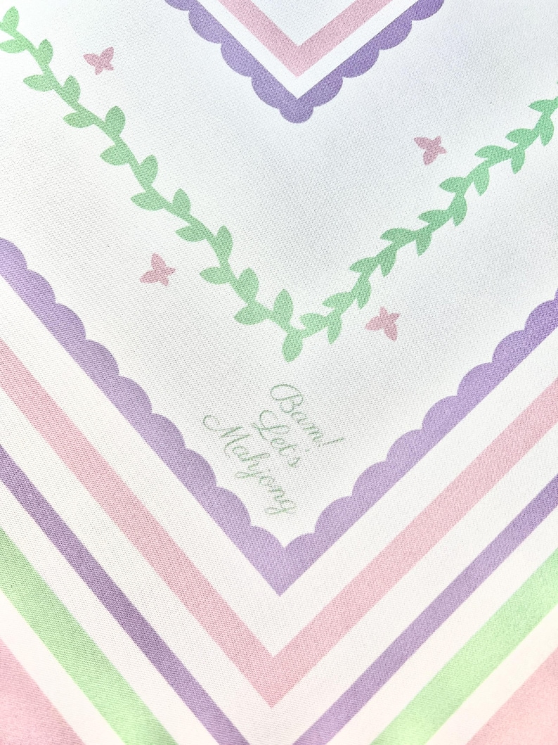 Op de afbeelding: Een witte textiel met een herhalend patroon van lavendel, roze en groene strepen en geschulpte randen. Het ontwerp bevat groene bladranken, roze vlinders en de tekst "Bam! Let's Mahjong" in lichtgrijs.