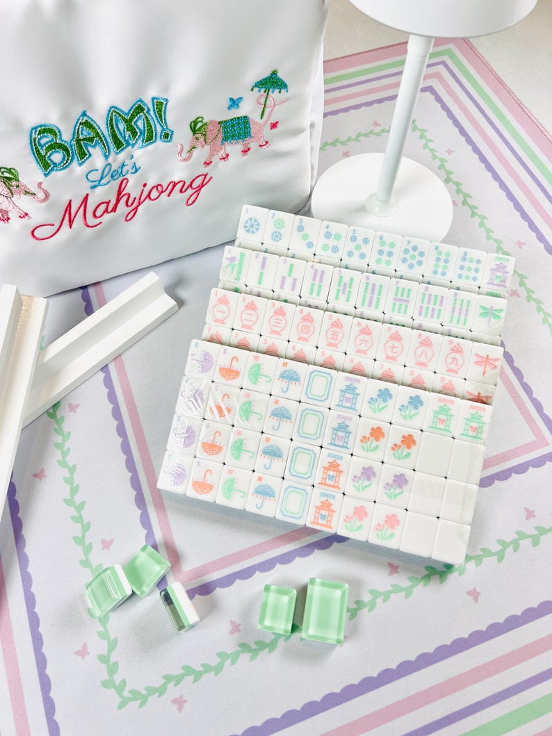 May include: A white satin bag embroidered with the words "BAM! Let's Mahjong" and an elephant design. Several stacks of white Mahjong tiles with colorful symbols and a few green translucent game pieces are arranged on a patterned surface.