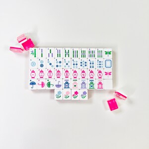 May include: A set of white Mahjong tiles with colorful symbols and characters, including bamboo, dots, and dragons. The tiles are arranged on a white surface, with several pink and white tiles scattered around. The game pieces feature blue, green, pink, and purple accents.